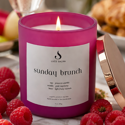 Pink candle labeled 'Sunday Brunch' on a table with raspberries and a bottle of wine.