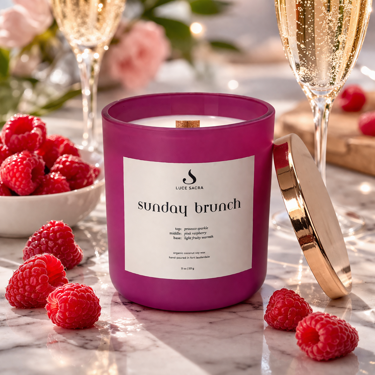 Fuscia candle labeled 'Sunday Brunch' with raspberries and champagne glasses on a marble surface.